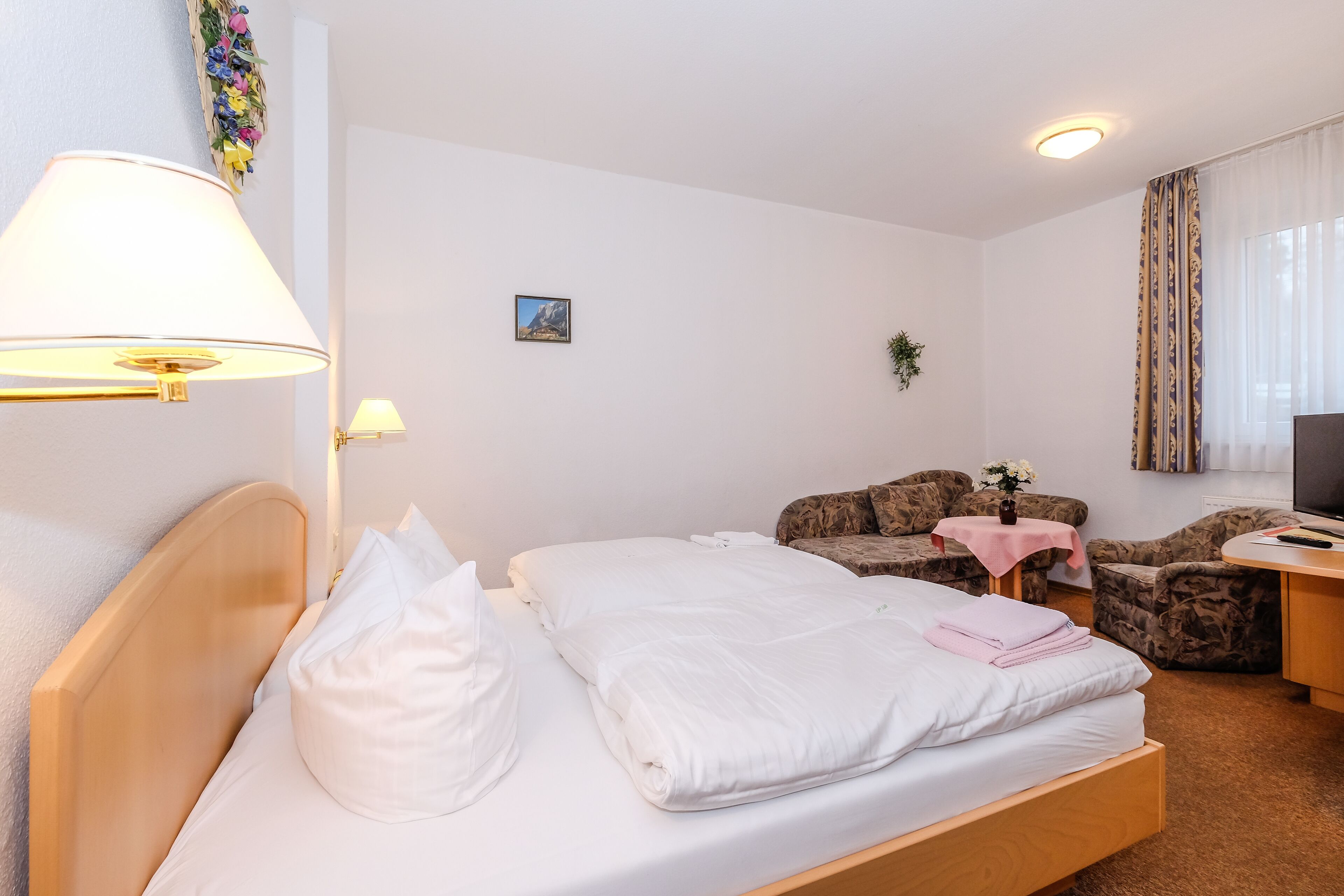 A hotel room with a bed, a desk, a chair, a small table, a lamp, and a TV.