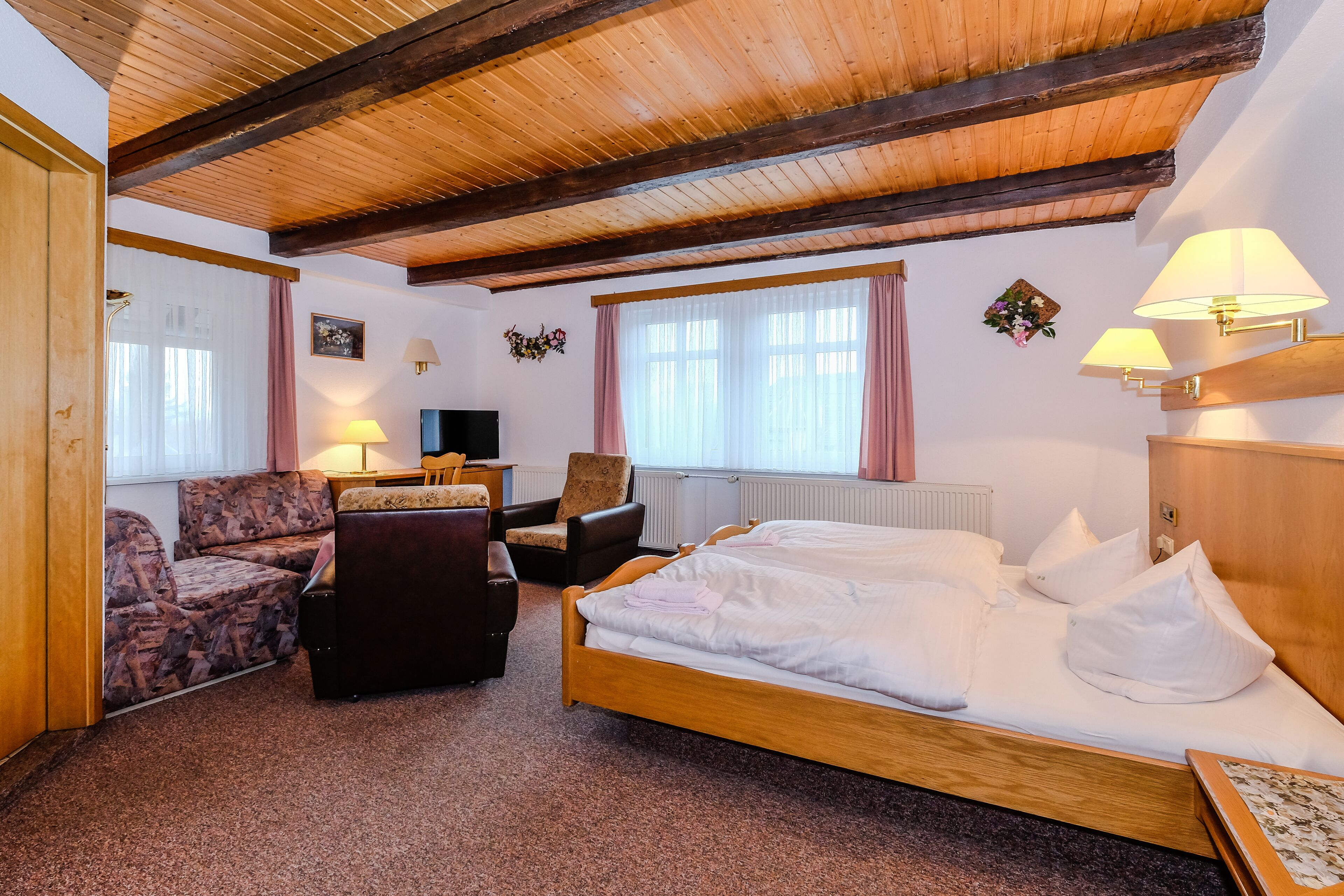 Habitación de hotel con techo de madera, cama, sofá, sillón, escritorio y dos lámparas.
