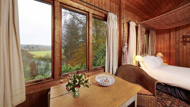 A wooden cabin room with a bed, a wicker chair, a table with a vase of flowers, and large windows offering a view of the outdoors.