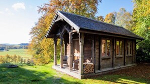 A rustic wooden cabin with a porch, a stack of firewood, and a view of the countryside.