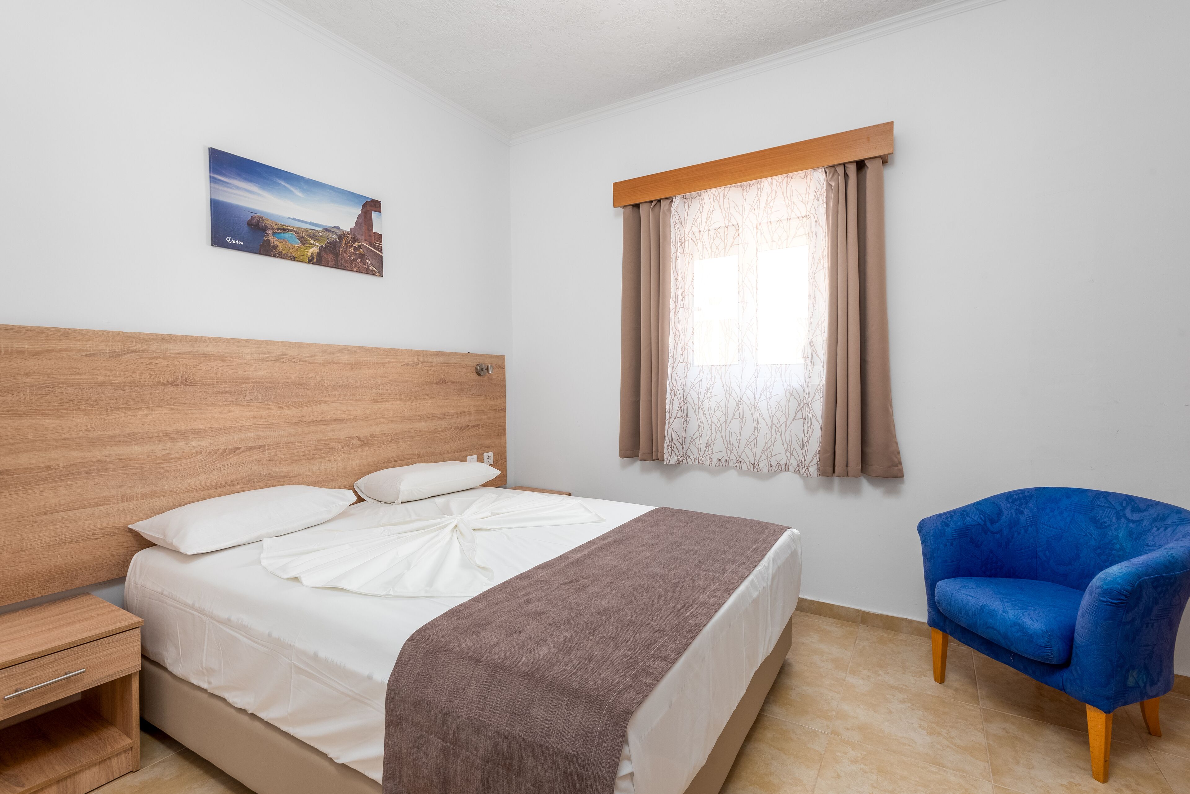 A bedroom with a wooden headboard, a bed with white linens, a brown throw blanket, a blue armchair, and a window with curtains.