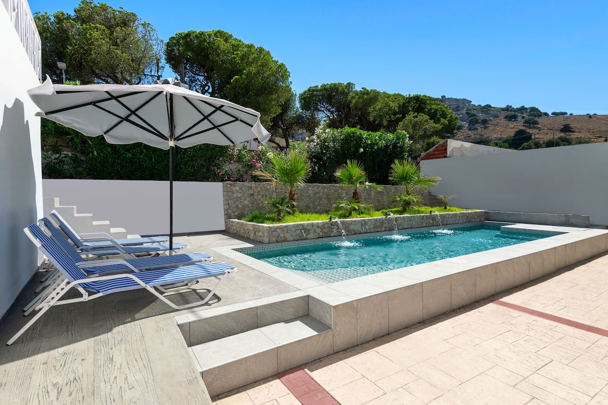 Una piscina con borde de piedra, sillas de descanso azules y una sombrilla.