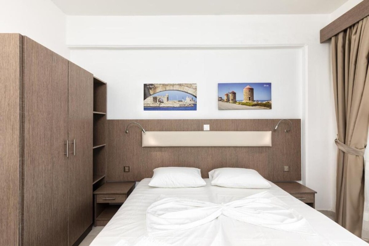 A hotel room with a bed, a wardrobe, a bedside table, and two framed pictures on the wall.