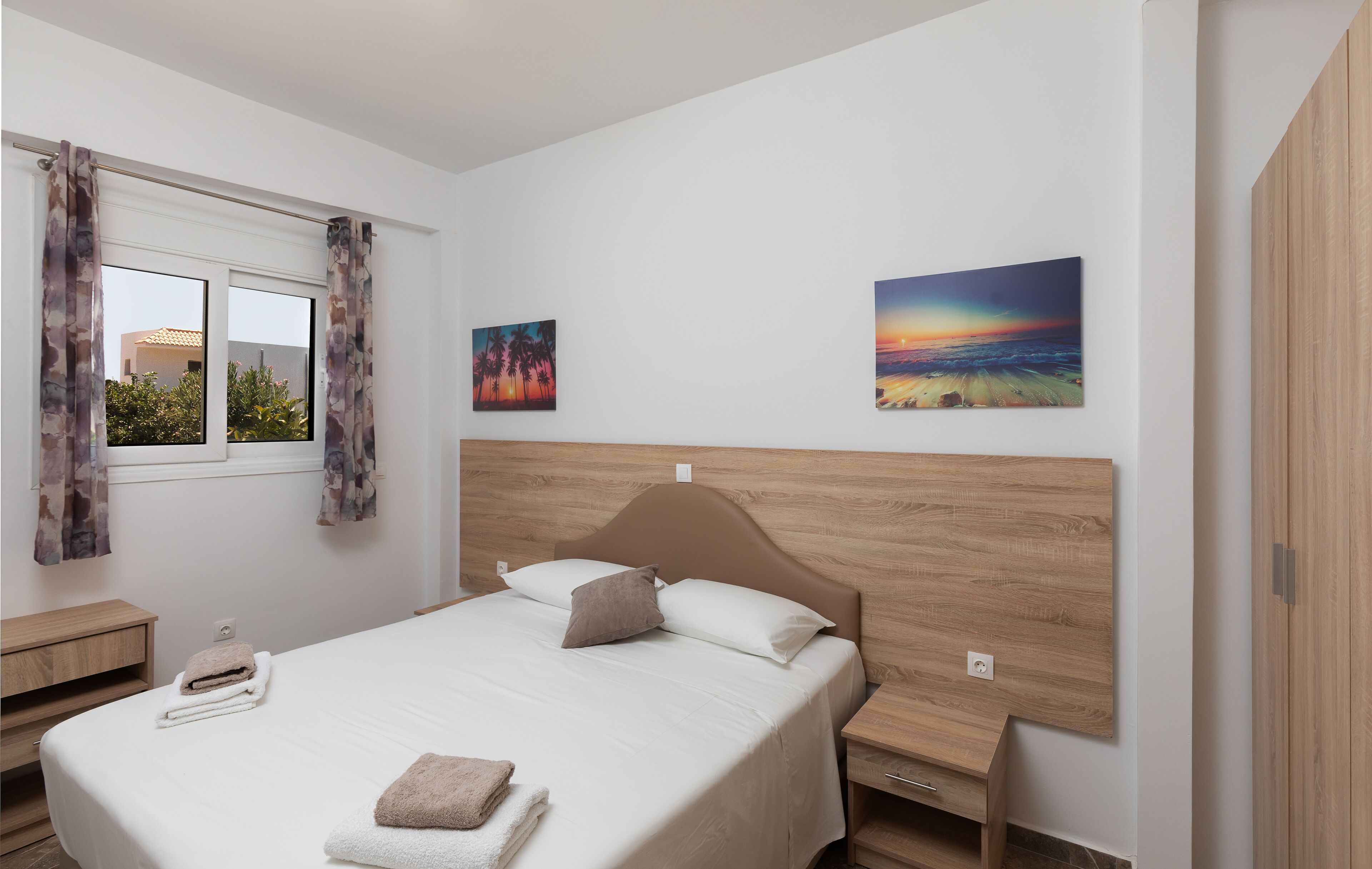 A modern bedroom with a wooden headboard, a large bed with white linens, two bedside tables, and two wall-mounted paintings.