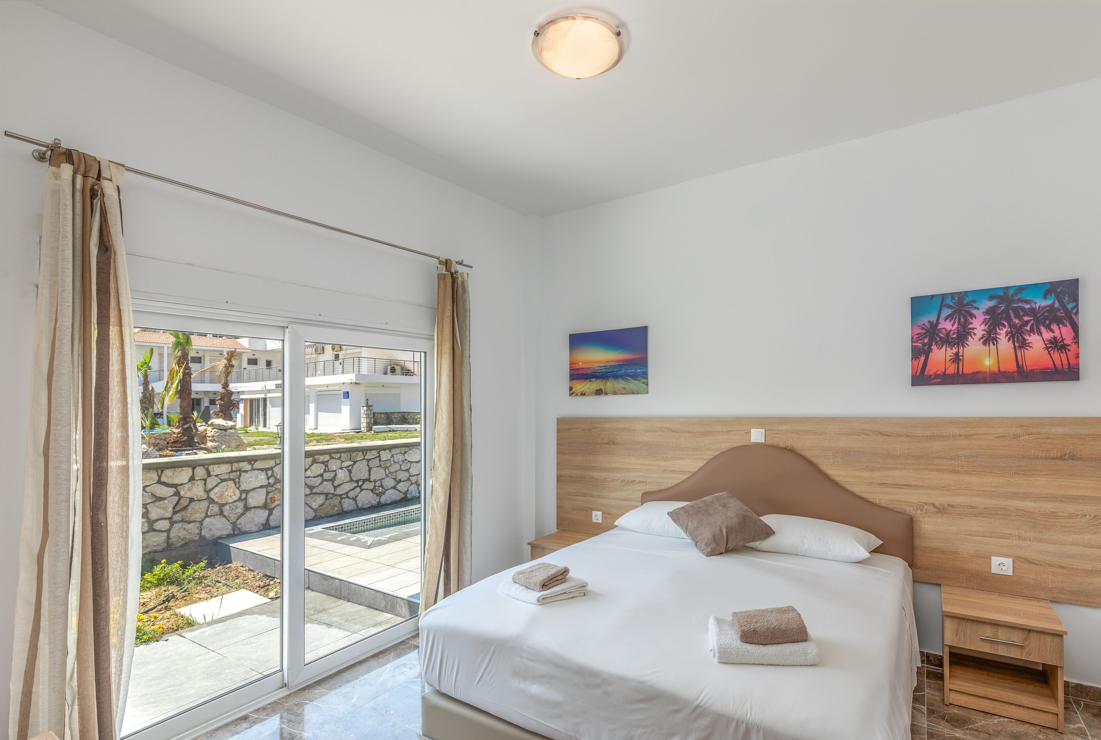 A bedroom with a large bed, a wooden headboard, a nightstand, and a view of a balcony with a stone wall and palm trees.