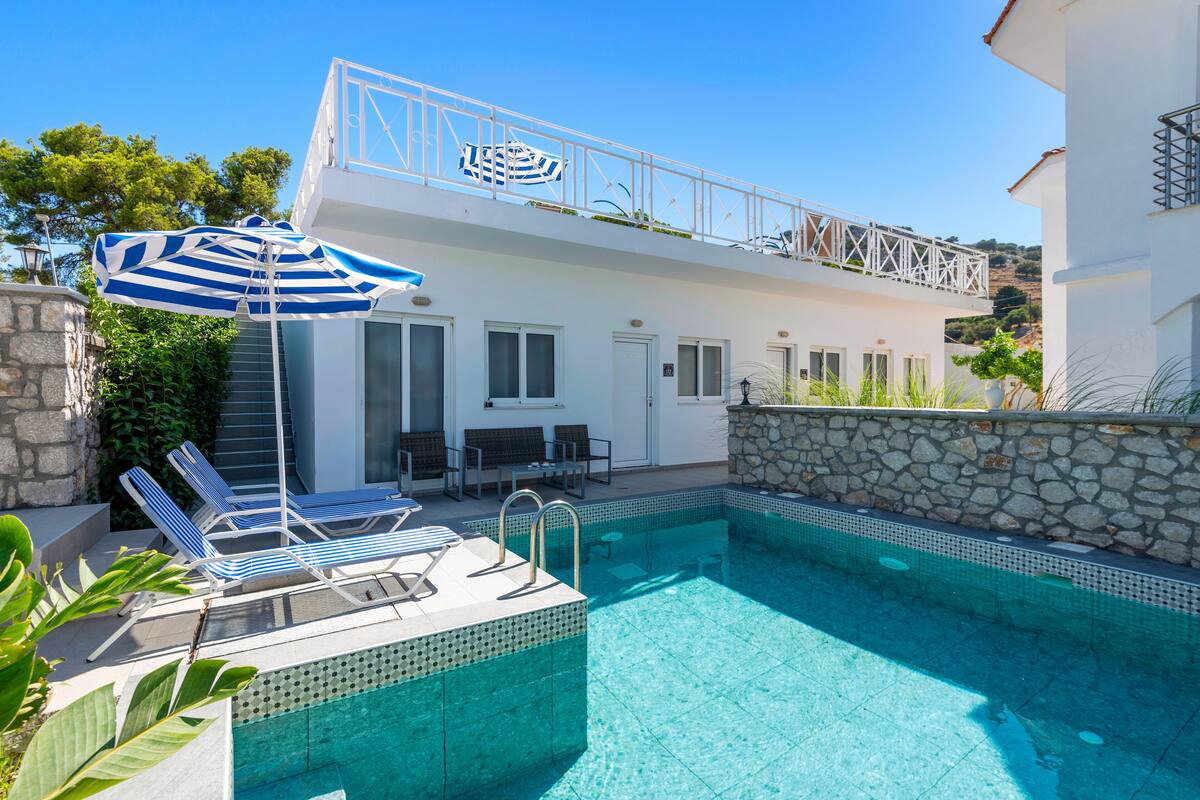 Un área junto a la piscina con sillones reclinables, una sombrilla a rayas y un edificio blanco con balcón.