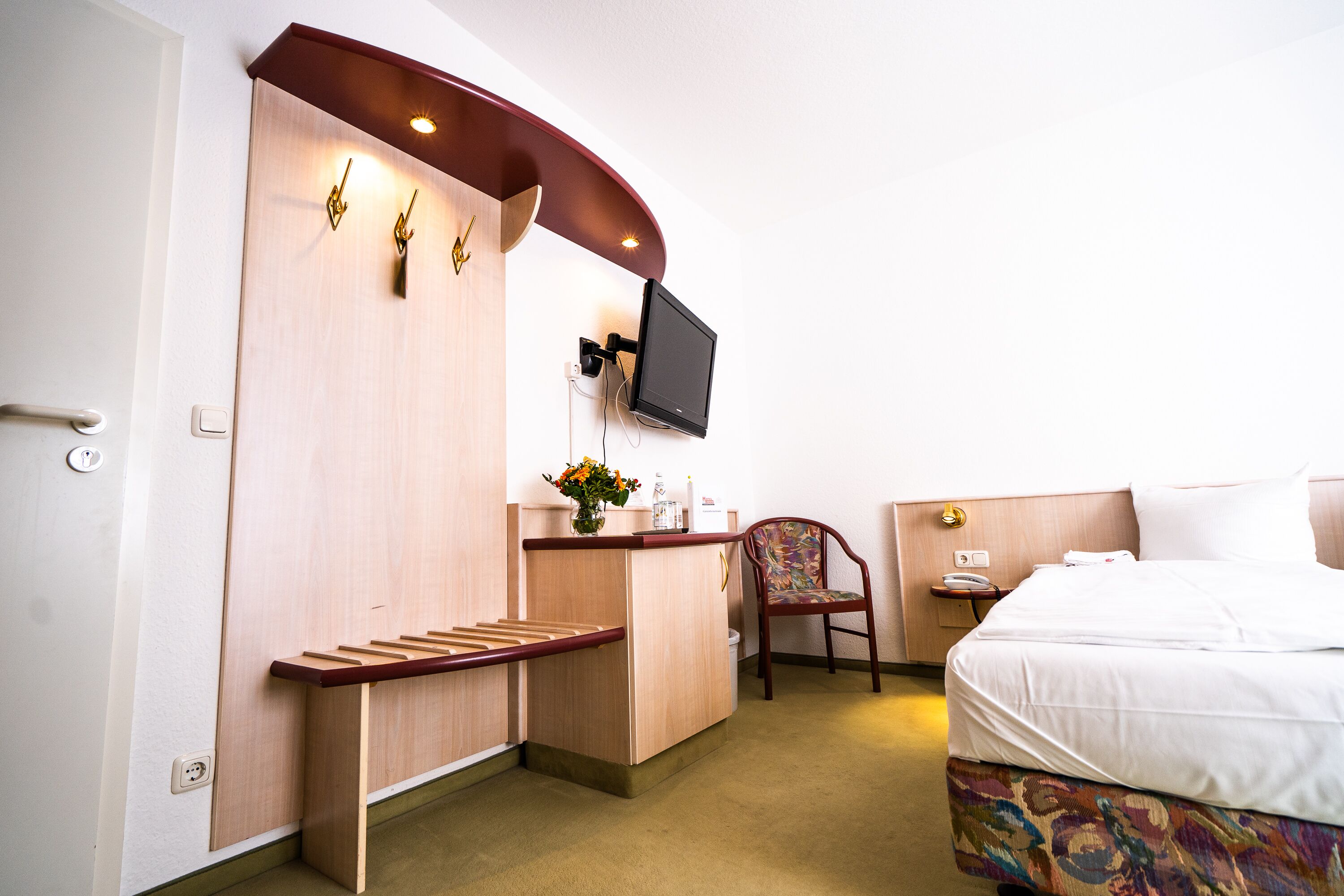 A hotel room with a bed, a desk with a chair, a TV mounted on the wall, and a small table with a vase of flowers.