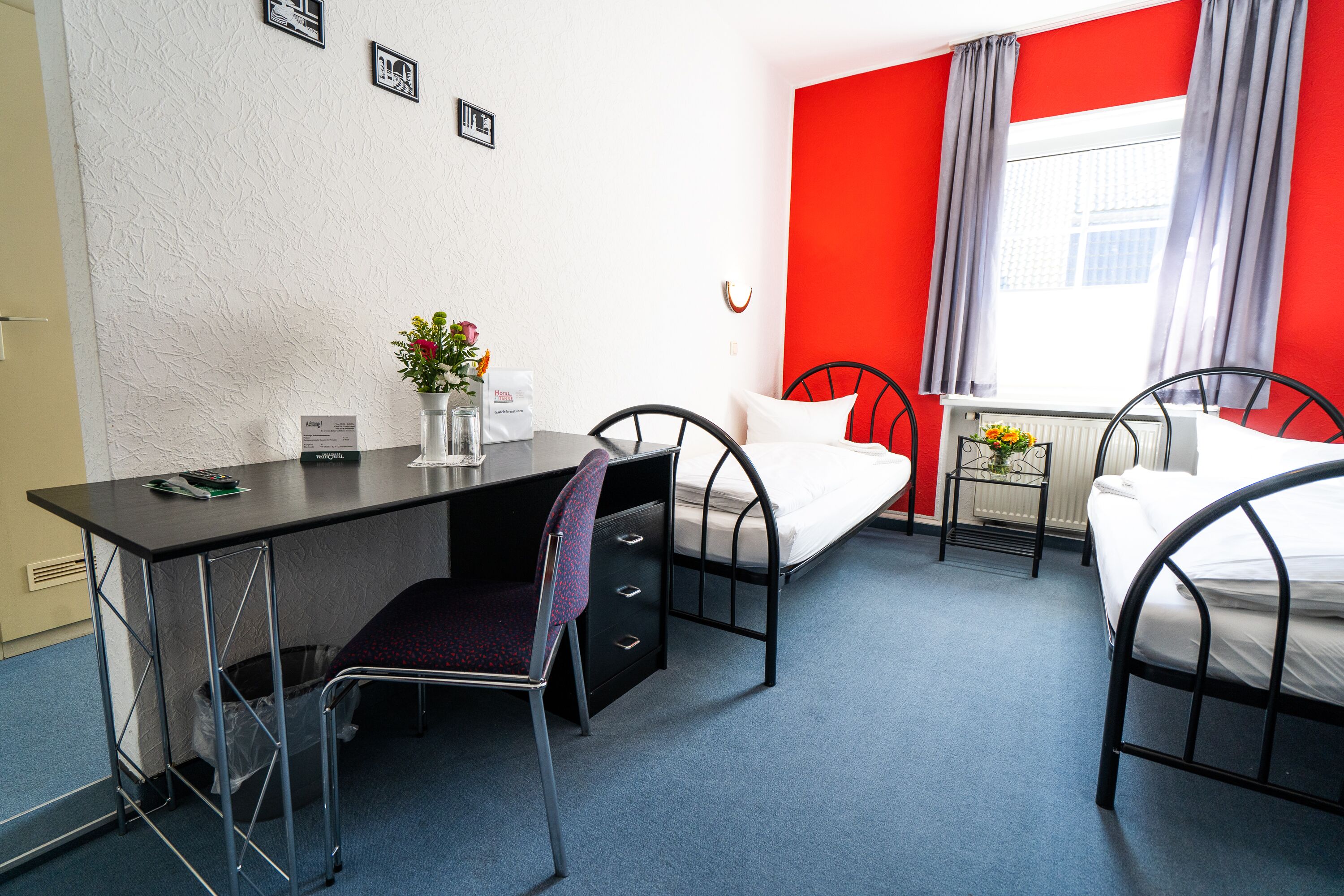 A hotel room with a desk, two single beds, a chair, a radiator, and a window with curtains.