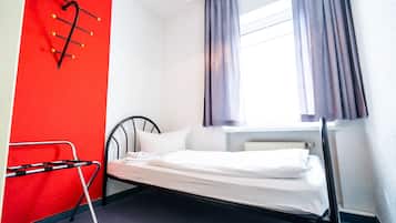 A single bed in a room with a red wall, a window with curtains, and a radiator.