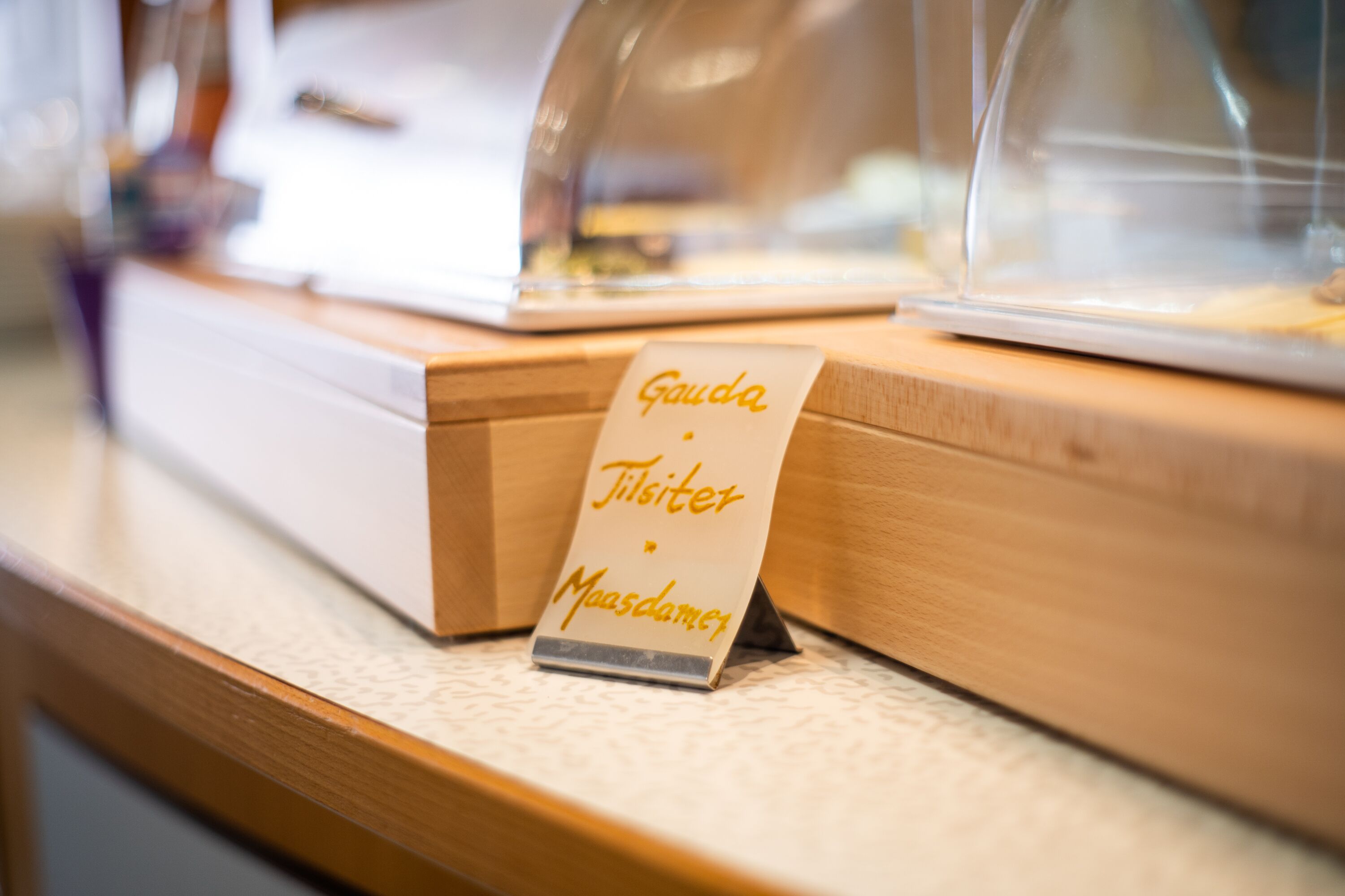 A sign with the text "Gauda Tilsiter Maasdammen" on a wooden counter.