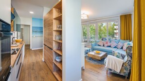 Cozinha moderna com prateleiras de madeira, área de refeições com sofá azul e vista para o mar através de grandes janelas.