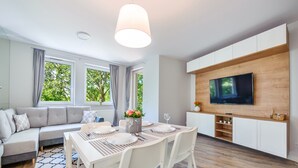 Uma sala de estar moderna com área de jantar, um sofá, uma televisão fixada numa parede de madeira e grandes janelas com cortinas.