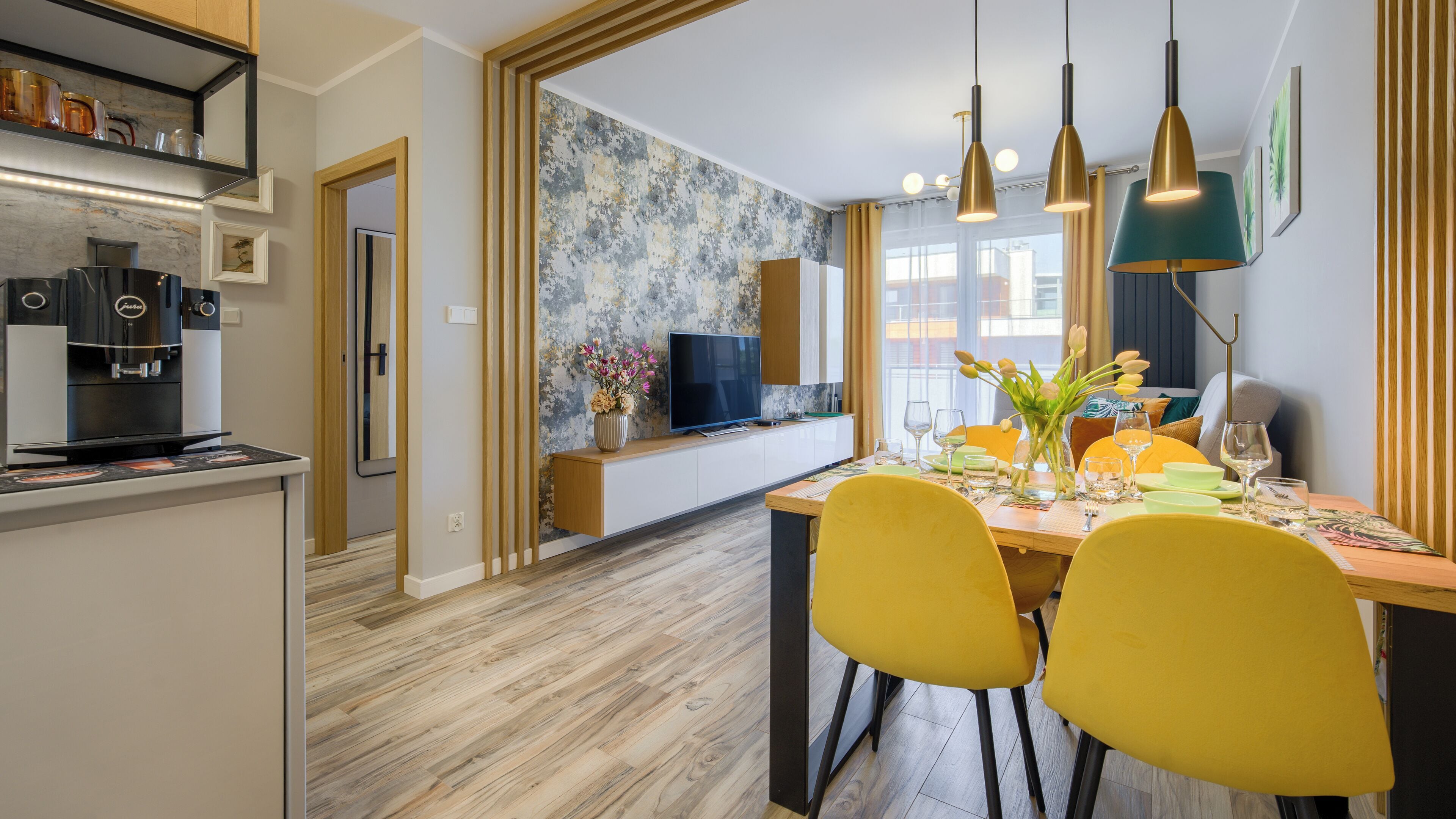 A modern kitchen with a coffee machine, a dining area with yellow chairs, a TV on a low wall, and a large window.