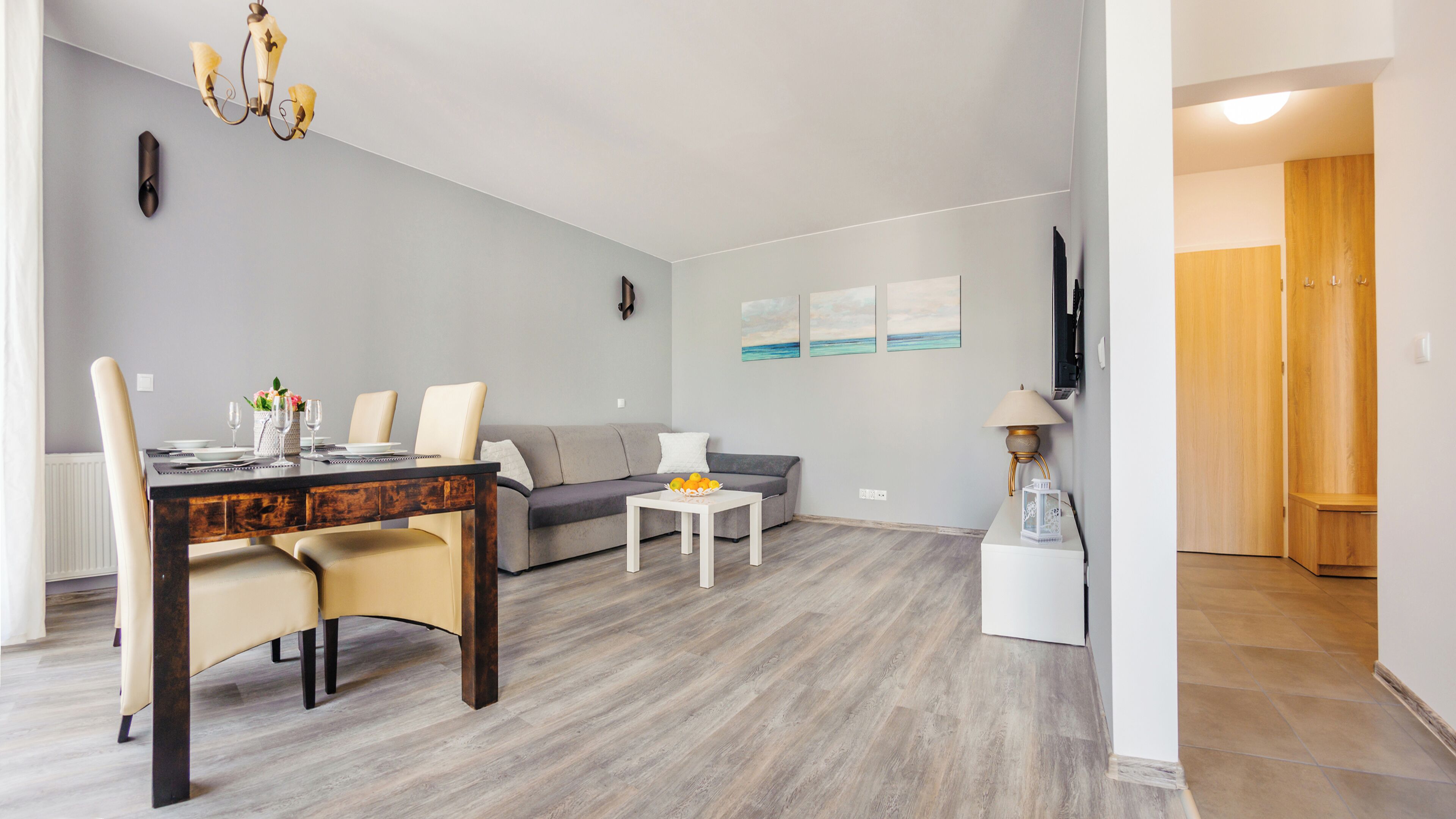 A modern living room with a dining area, grey sofa, wooden dining table, and a small white side table.