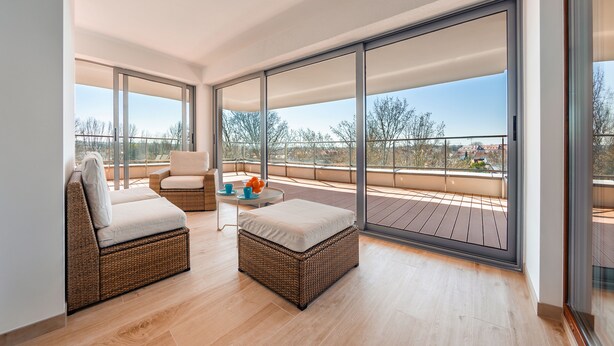 Uma varanda espaçosa com um sofá e poltrona de vime, uma mesa pequena com almofada e vista para a cidade.