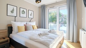 Quarto bem iluminado com uma cama grande, várias almofadas, uma mesa de cabeceira e vista para a paisagem urbana através de uma janela ampla.