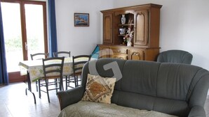 A living room with a grey sofa, a wooden cabinet, a dining table with chairs, and a blue curtain. - Cozy Forest-Edge Vacation Home for 4 near Jard-sur-Mer (Saint-Vincent-sur-Jard)