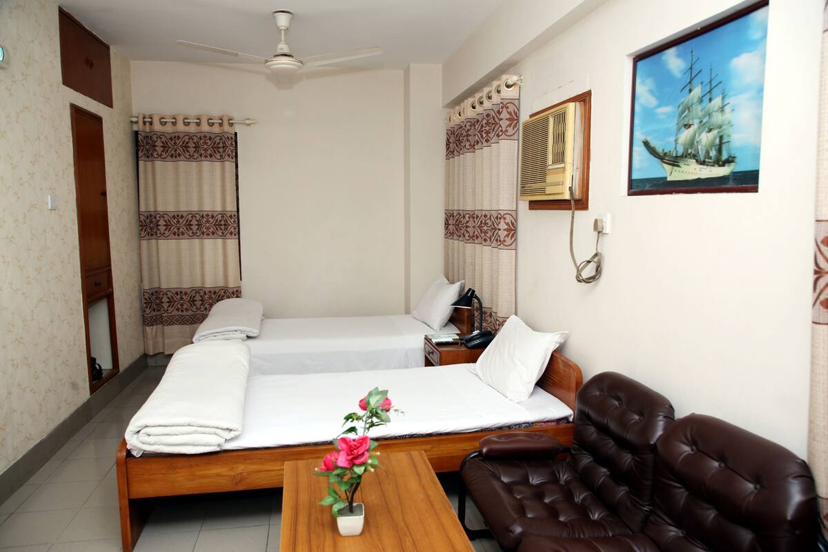 A hotel room with two beds, a wooden table with a vase of flowers, a brown leather chair, a ceiling fan, and a painting of a ship on the wall.
