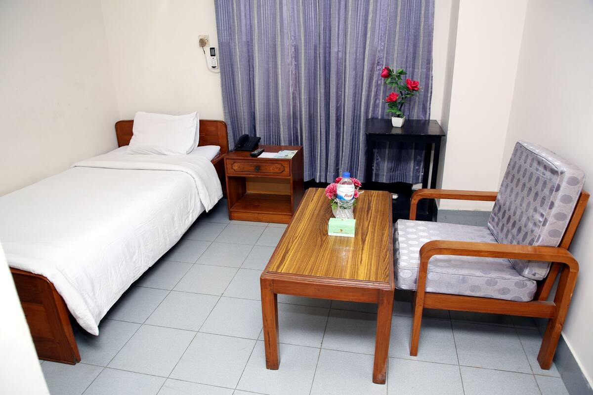 A hotel room with a single bed, a wooden nightstand, a wooden table, a grey patterned armchair, and a small table with a vase of red flowers.