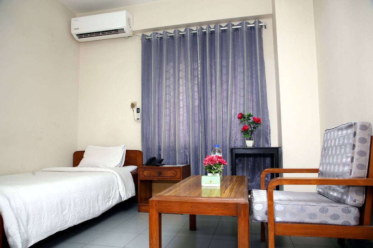 A hotel room with a bed, a wooden table, a chair, a nightstand, a lamp, and a curtain.