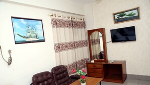Ein Zimmer mit brauner Ledersofa, einem Holztisch mit Blumenvase, einem Holzschrank, einem Flachbildfernseher, einem gerahmten Bild eines Schiffes und einem Vorhang mit Blumenmuster.