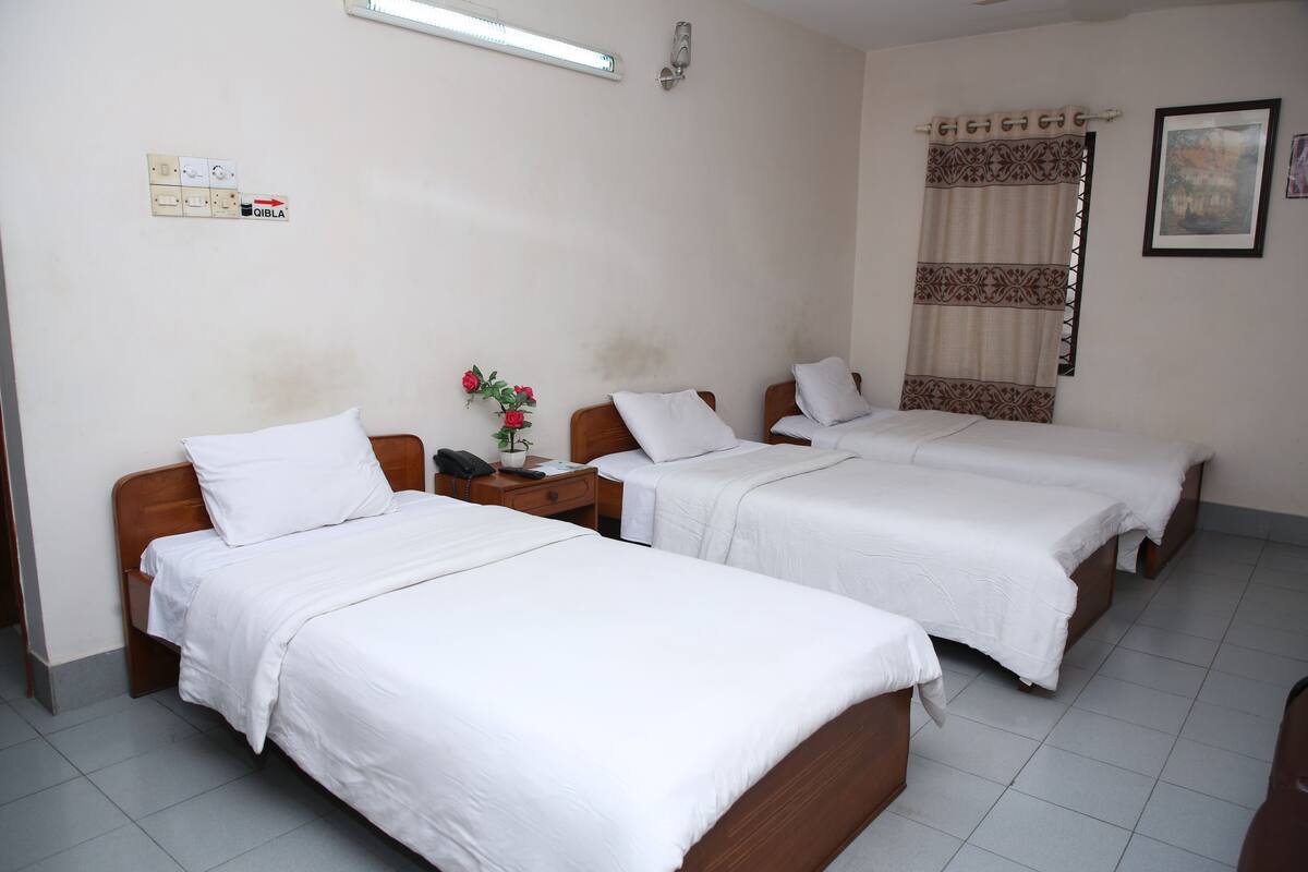 A hotel room with three single beds, a bedside table with a lamp, a small table with a vase of flowers, and a wall-mounted telephone.