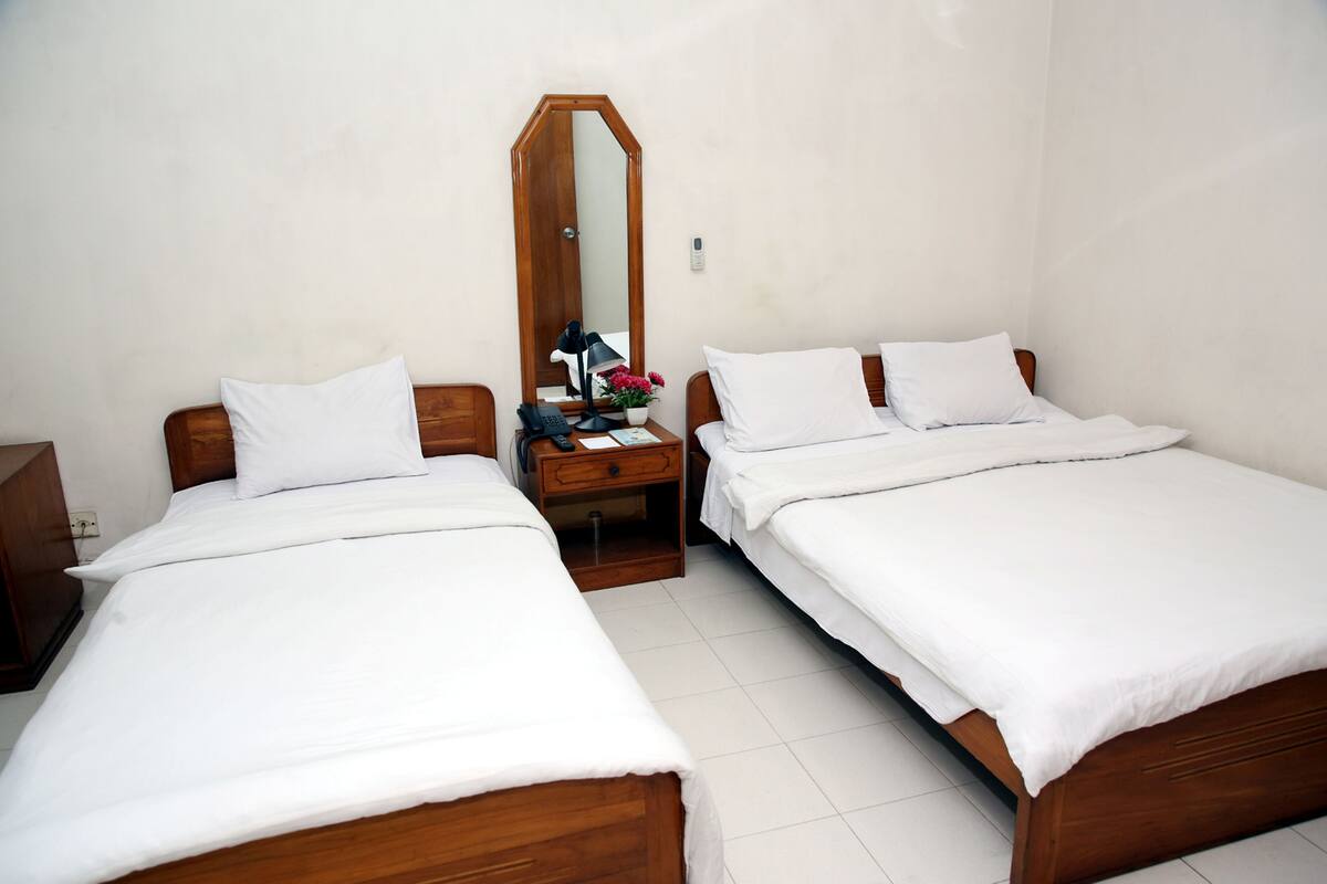 A hotel room with two single beds, a wooden nightstand with a lamp, a mirror, and a small table with a phone and flowers.