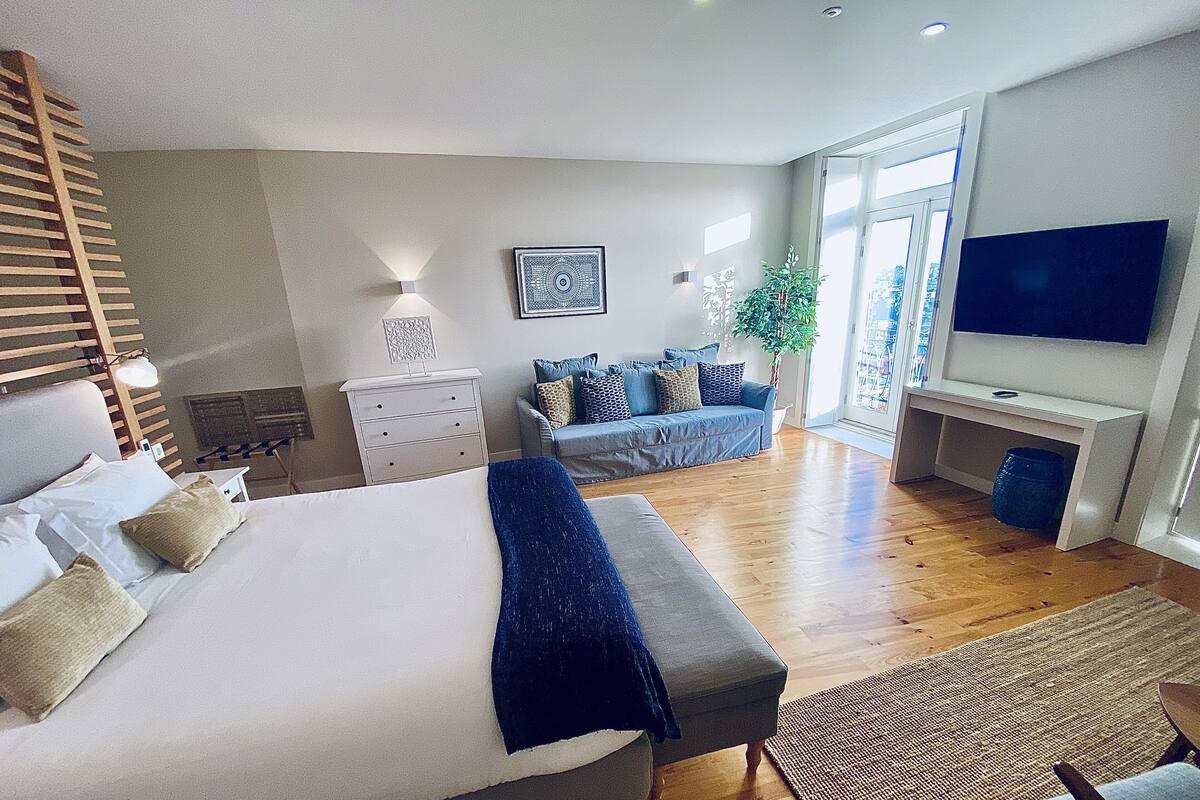 A modern bedroom with a large bed, bedside tables, a seating area with a sofa, a TV, and a large window.