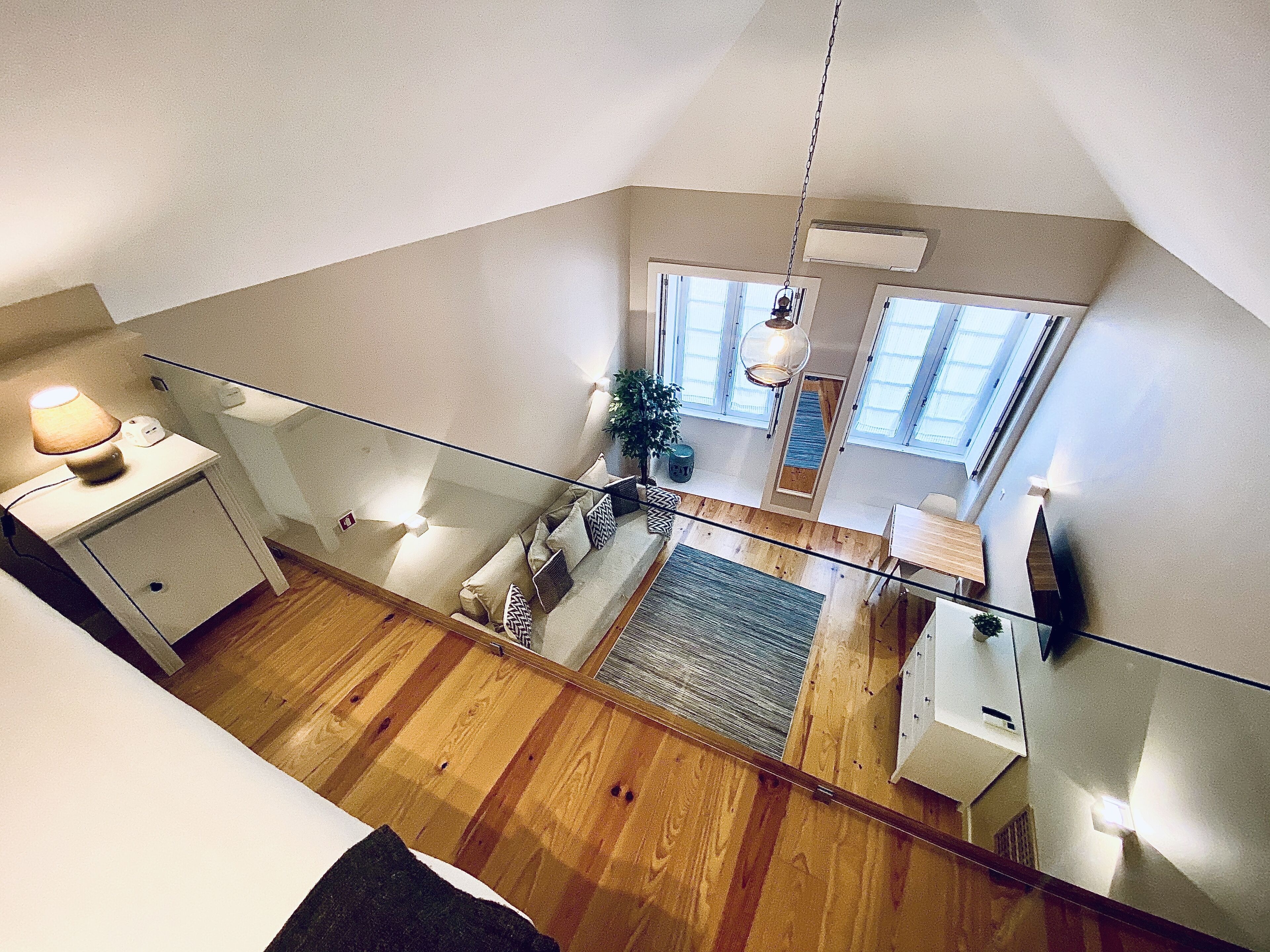 A high-ceilinged room with a glass railing, wooden flooring, a sofa, a table, and a hanging light fixture.