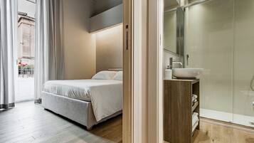 Una habitación de hotel moderna con una cama, una mesita de noche de madera, un box de ducha de vidrio y vistas a la calle.