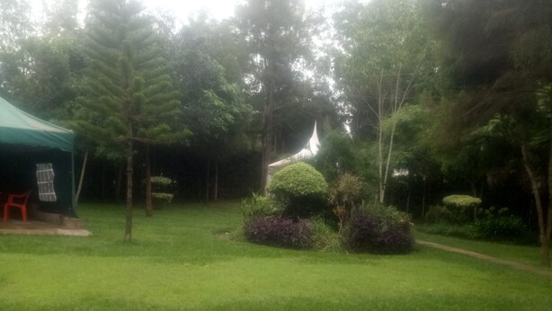 A well-maintained grassy area with a pathway, a small structure with a green roof, and various plants and trees. - Reed Hotel Annex (Ongata Rongai)