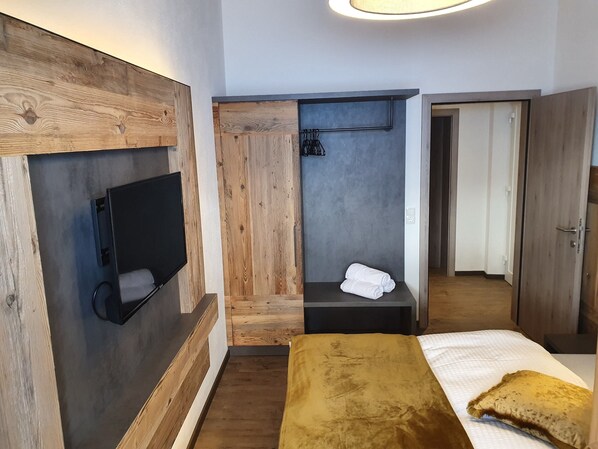 A modern hotel room with a wooden accent wall, a flat-screen TV, a built-in shower, and a bed with a mustard-colored throw.