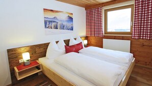 Un dormitorio de cabaña de madera con una cama, una mesita de noche, una lámpara de pared, una ventana con cortinas a cuadros y un cuadro de un paisaje montañoso nevado.