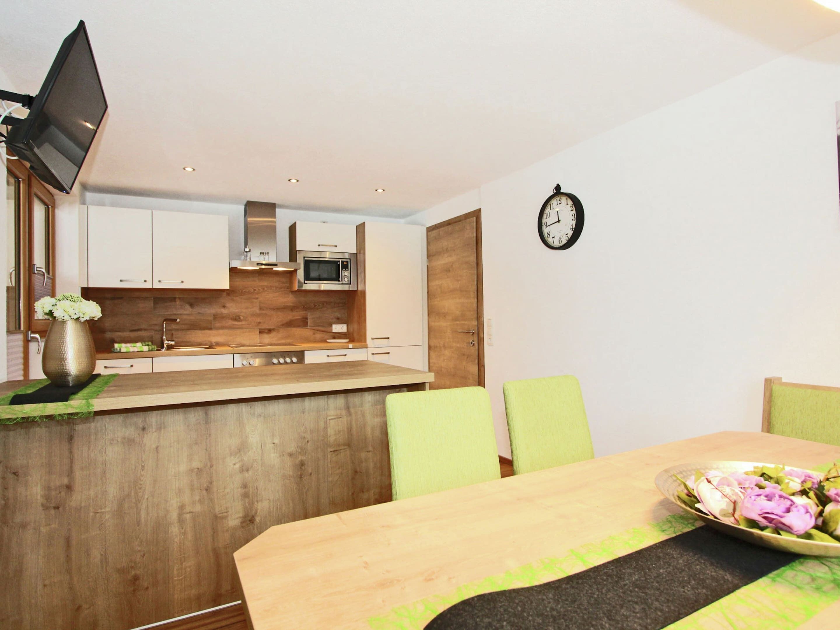A modern kitchen with white cabinets, a wooden countertop, and a built-in microwave. There is a dining table with green chairs and a clock on the wall.