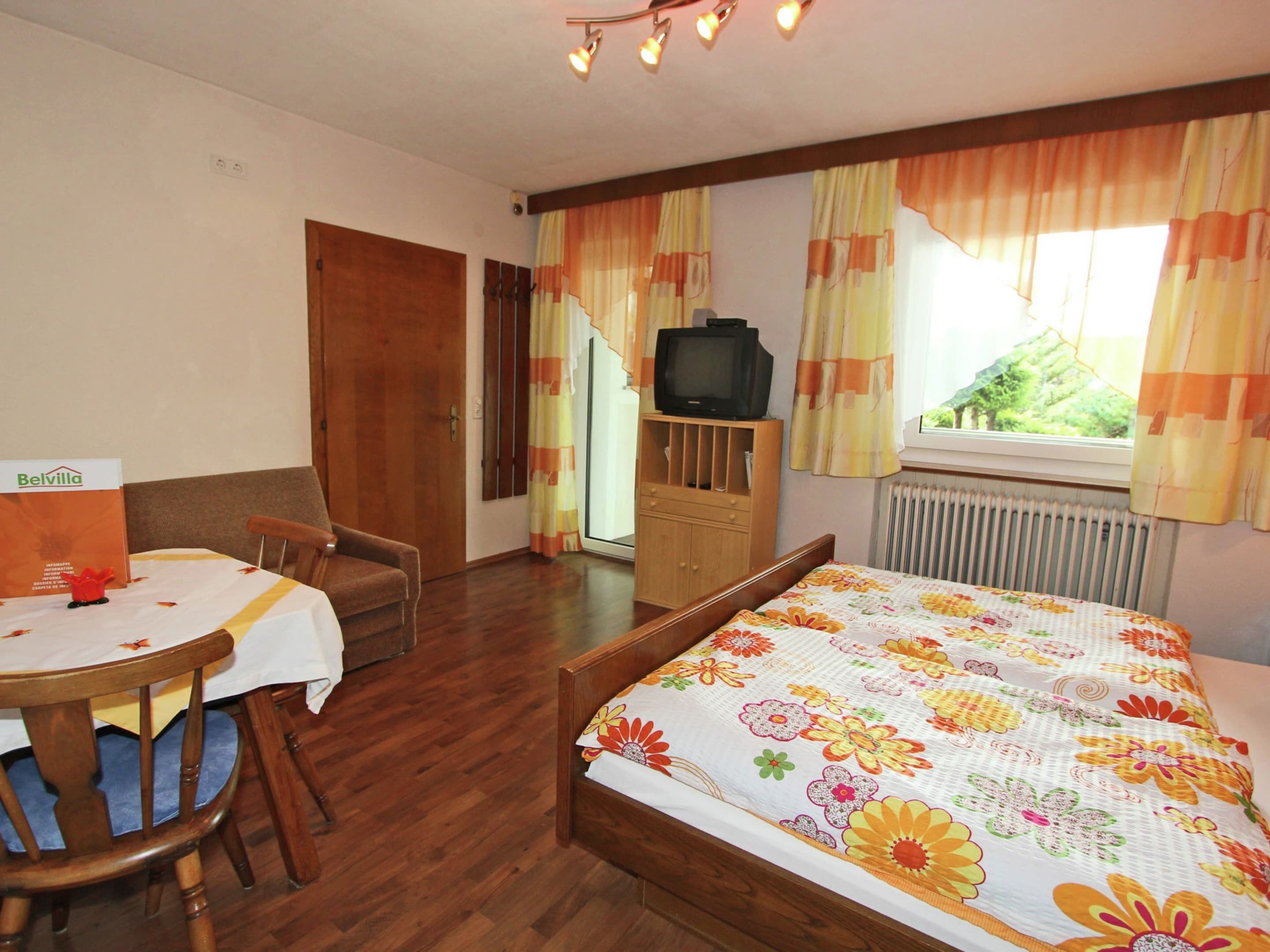 Une chambre à coucher avec un lit, une chaise en bois, une table avec une boîte, un téléviseur posé sur un meuble en bois et des rideaux.