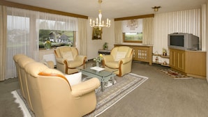 A living room with two beige armchairs, a glass coffee table, a chandelier, a television on a wooden cabinet, and a view of a house through the window. - Heavenly Apartment in Wängle Tyrol near Walking Trails (Waengle)