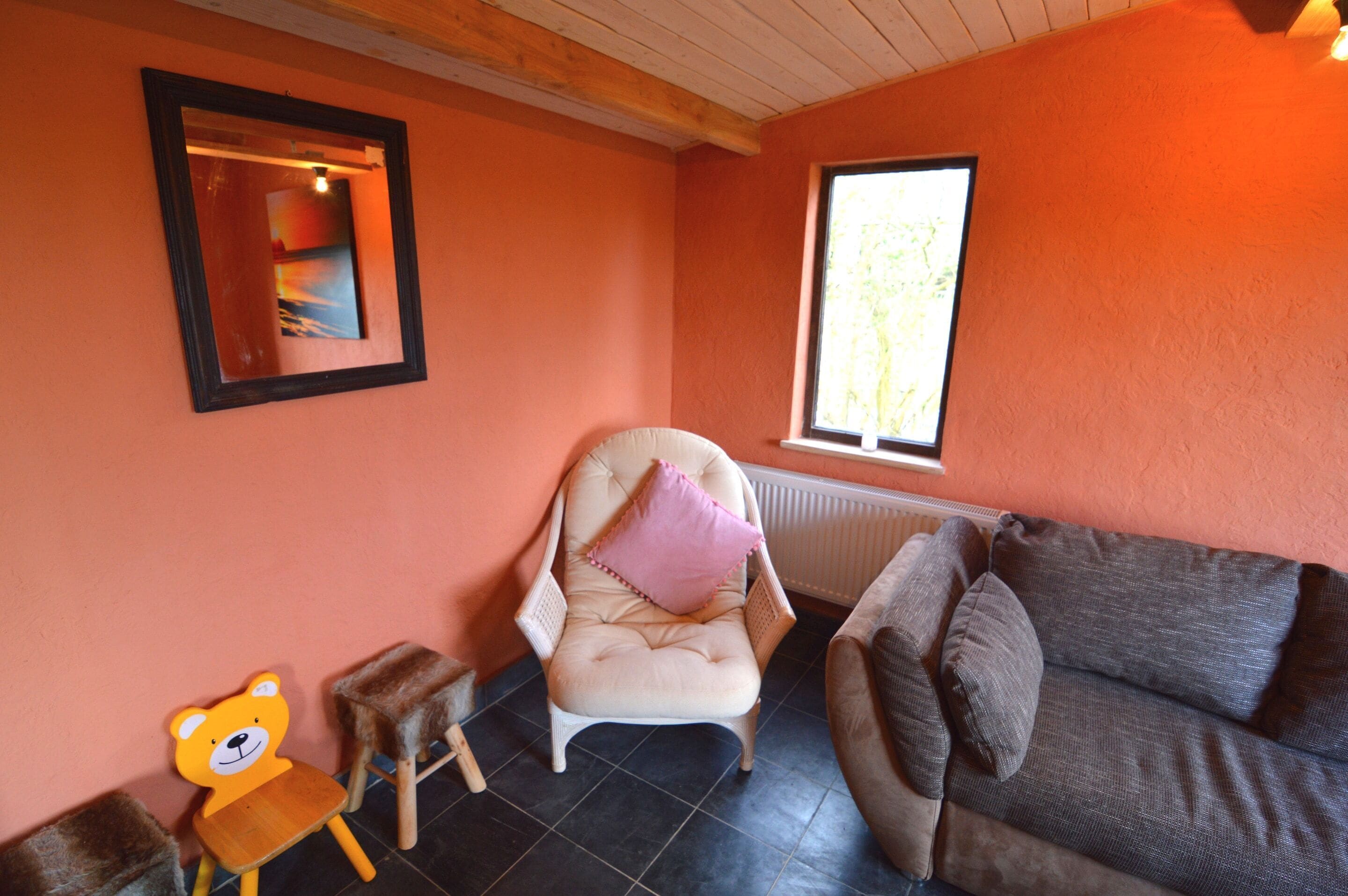 A room with a cushioned chair, a sofa, a small wooden stool, and a teddy bear.