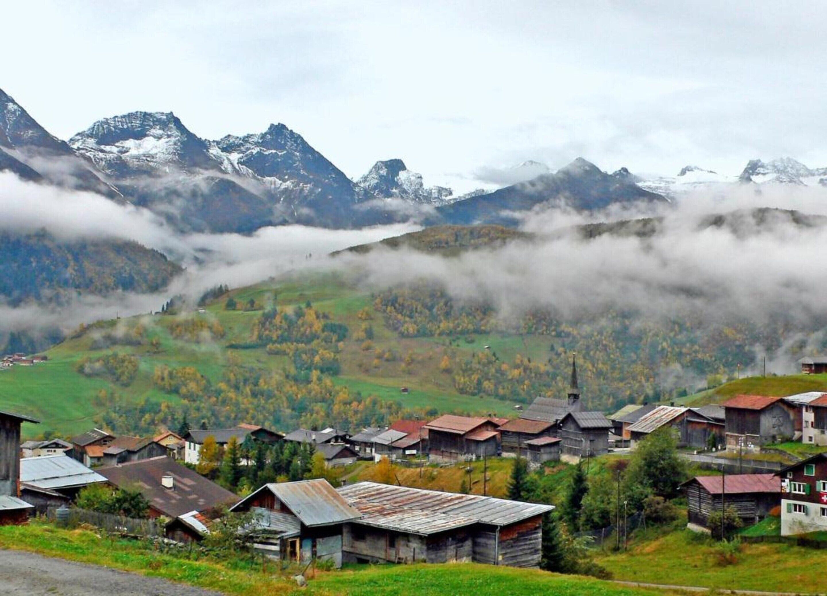 A rural village with traditional wooden houses, surrounded by mountains and covered by clouds.