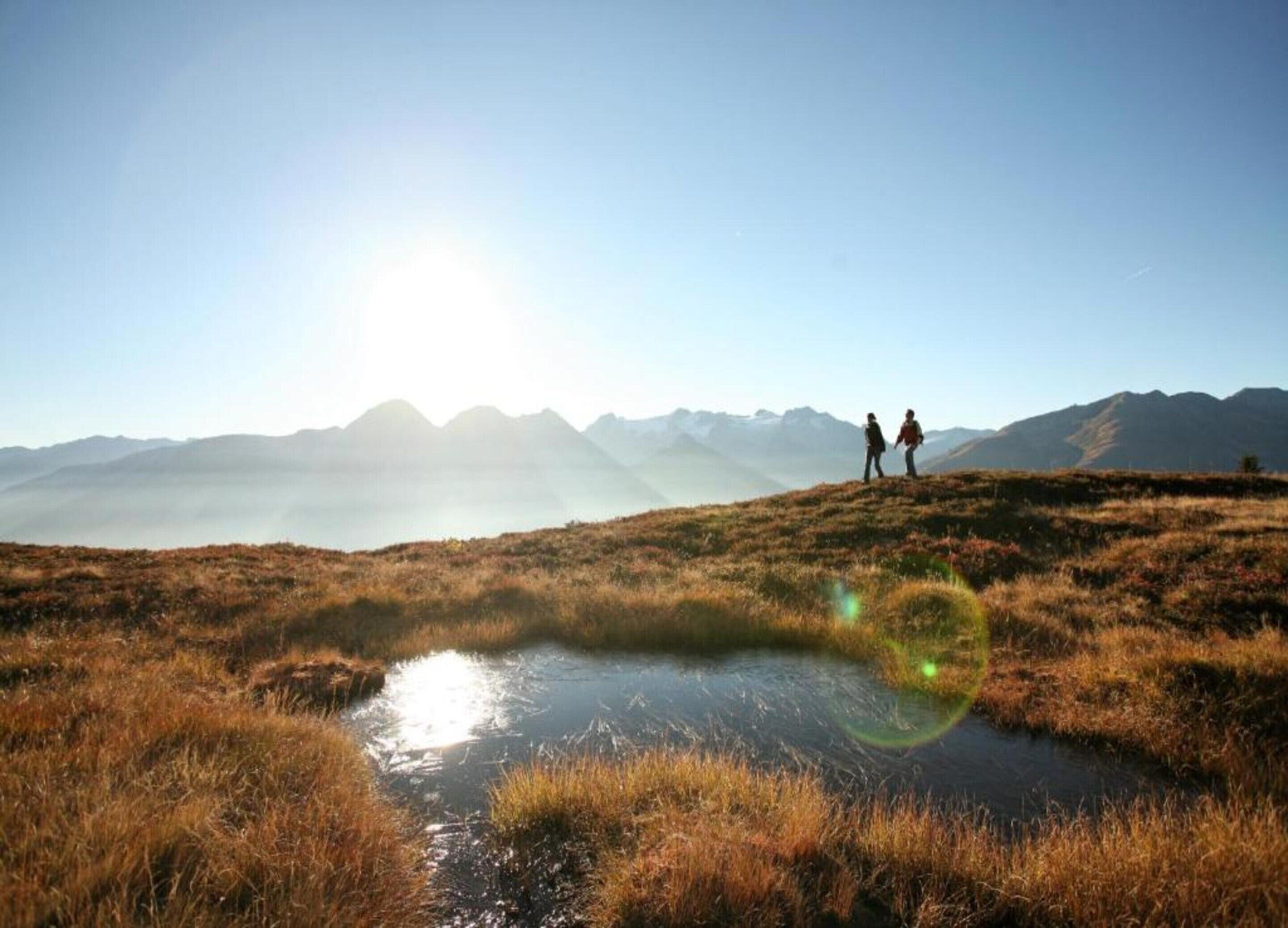 Ett naturligt landskap med en liten vattenyta, två personer som står på en gräsklädd kulle och ett bergskedja i bakgrunden.