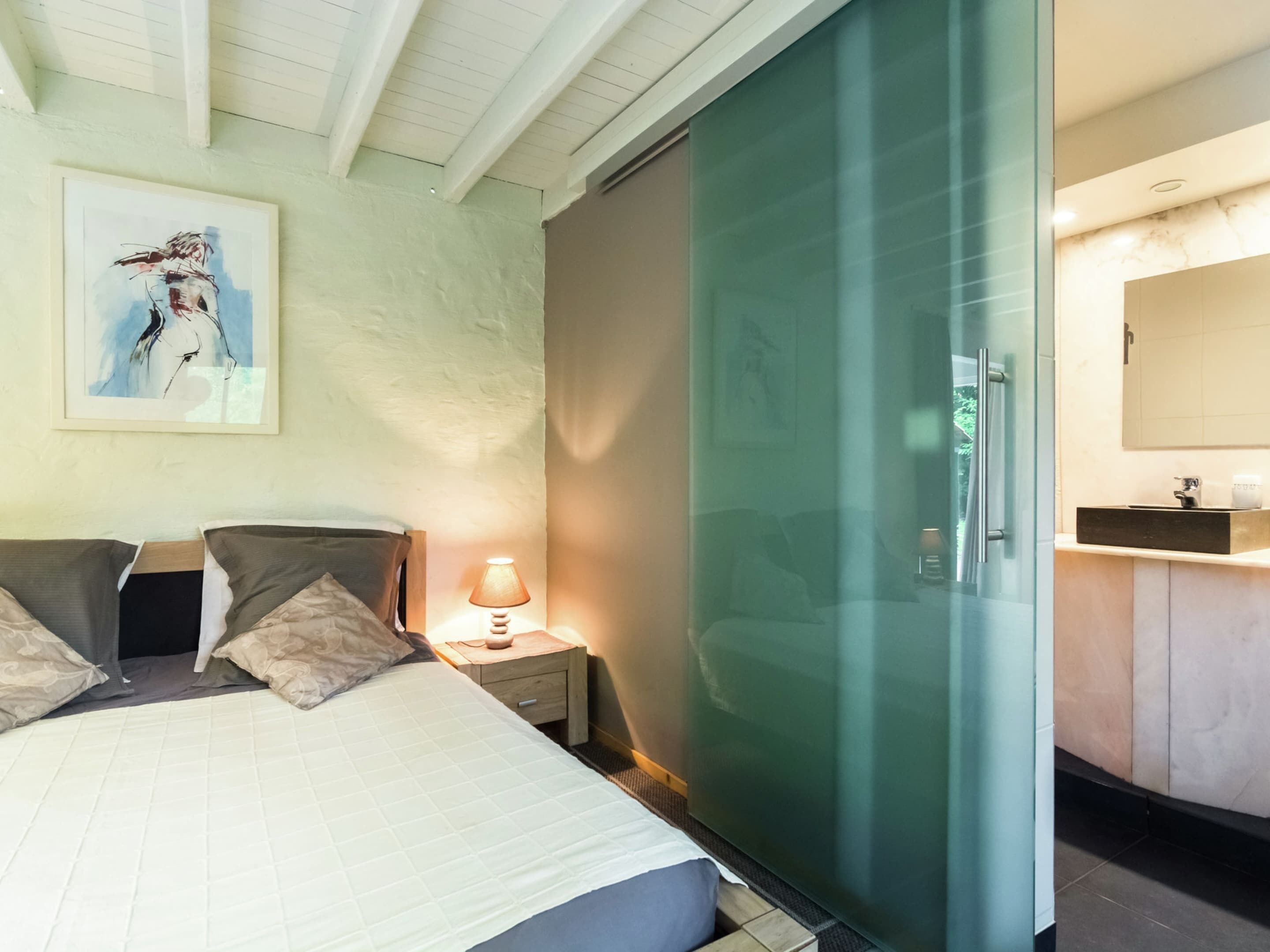 Un dormitorio con cama, mesita de noche, lámpara y una obra de arte en la pared.