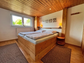 A wooden bed with two pillows, a window, a radiator, a lamp, and a small stool.