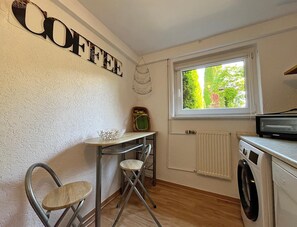 A compact kitchen with a washing machine, a radiator, a window with a view of greenery, a table with chairs, and a wall-mounted sign that reads 'COFFEE'.