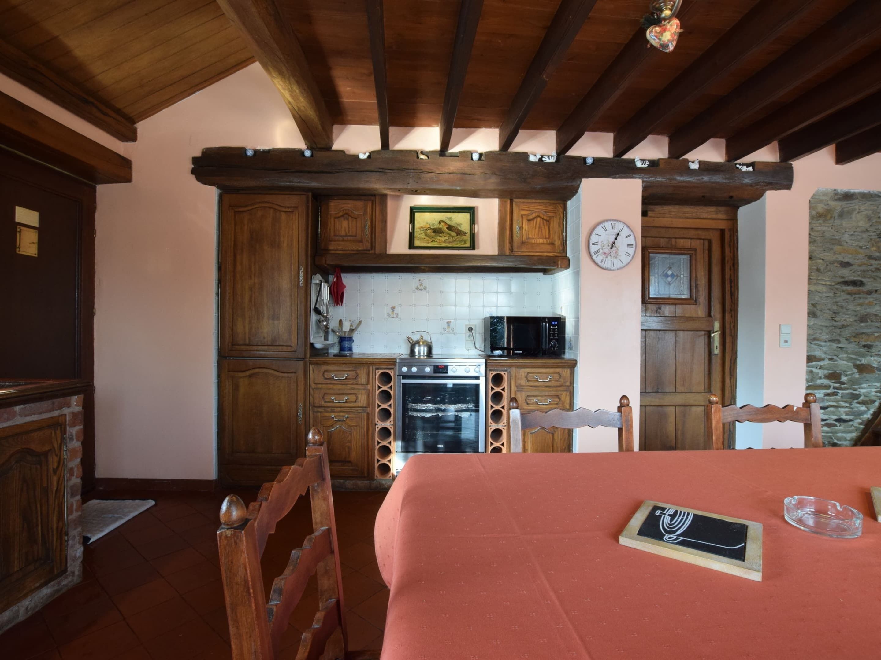 A rustic kitchen with wooden cabinets, a stove, and a microwave. There is a wooden chair and a bed with a red blanket.