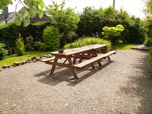 A wooden picnic table with benches in a garden area with gravel ground, surrounded by various plants and trees. - Holiday Home in Ronchampay (La Roche-en-Ardenne)