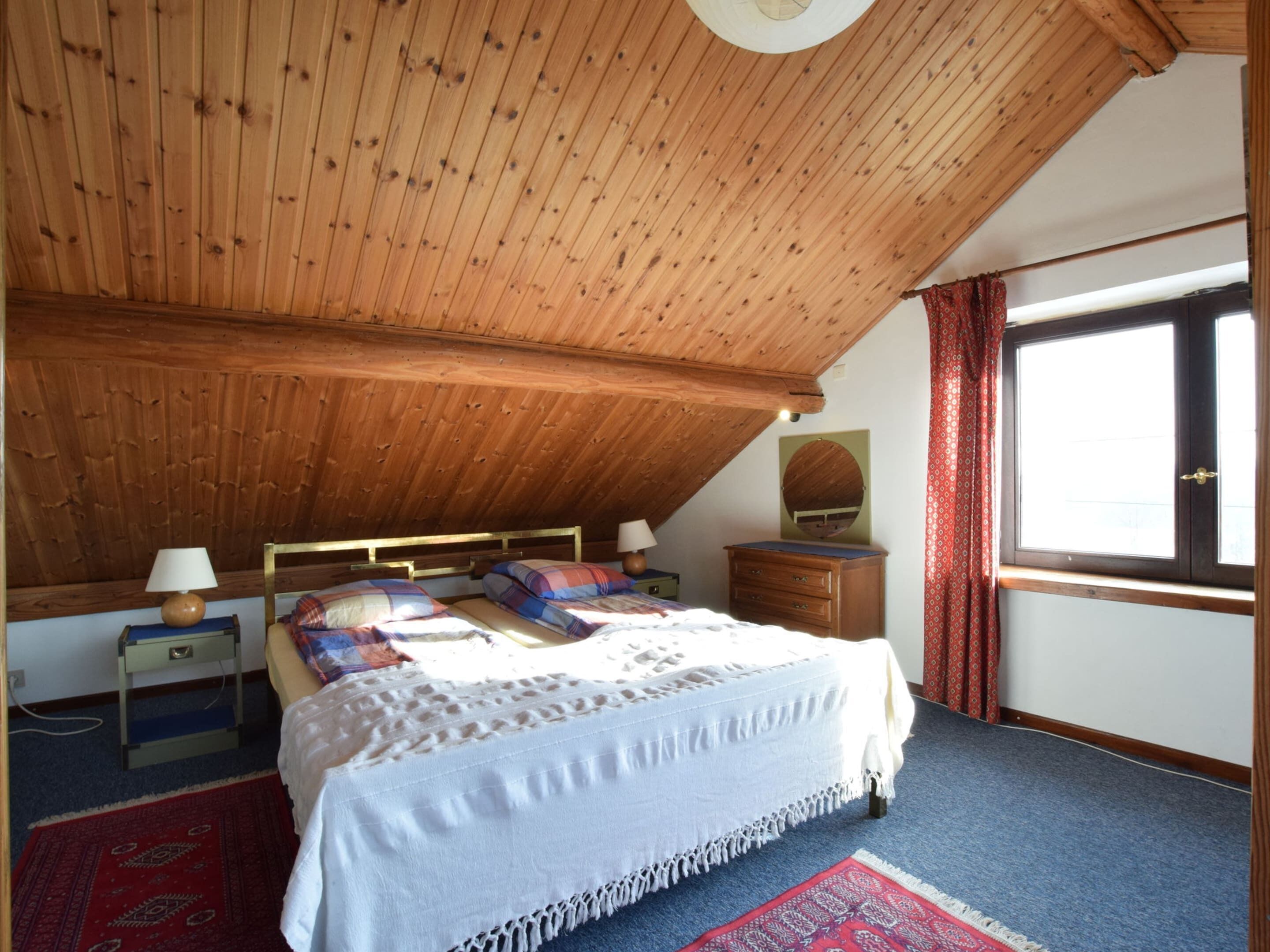 Ein Schlafzimmer mit Holzboden, einem Bett mit weißer Bettdecke, einem Nachttisch mit Lampe, einem Schminktisch und einem Fenster mit roten Vorhängen.