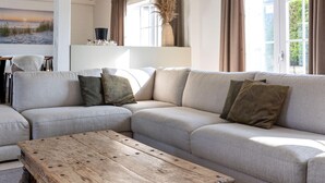 Ein modernes Wohnzimmer mit einem Ecksofa, einem Holztisch und einem großen Fenster mit Vorhängen.