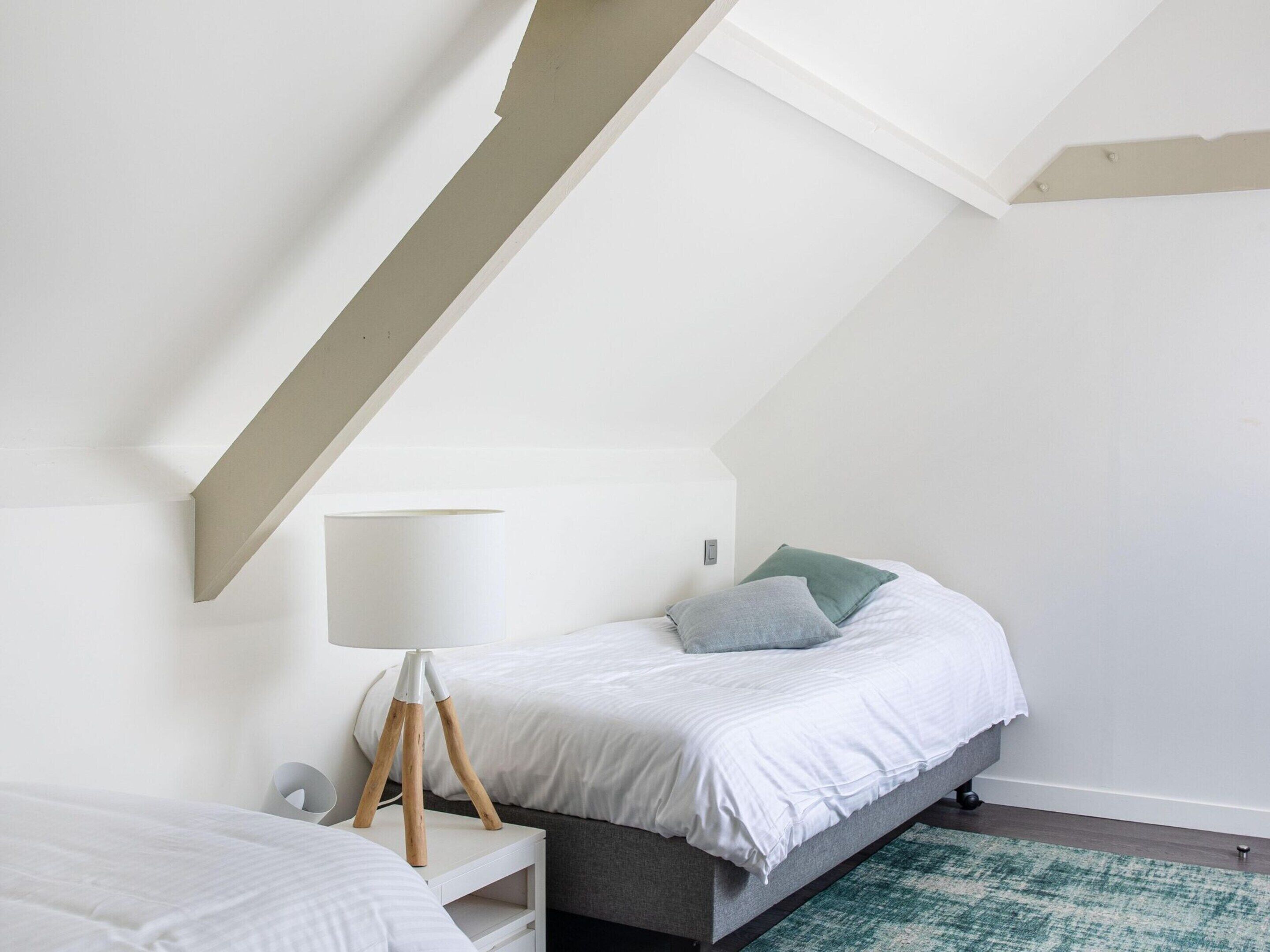 Un dormitorio con techo inclinado, una cama con ropa de cama blanca, una mesita de noche con lámpara y una alfombra verde.