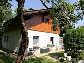 Et hus med skråtag, en lille veranda og en have med en stol og et bord.