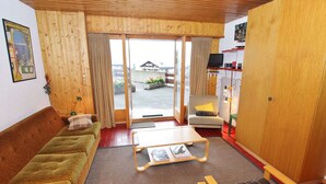A cozy room with a wooden ceiling, a green sofa, a brown sofa, a small table with magazines, and a view of the outside through a glass door. - Apartment Near the Ski Lifts (Riddes)