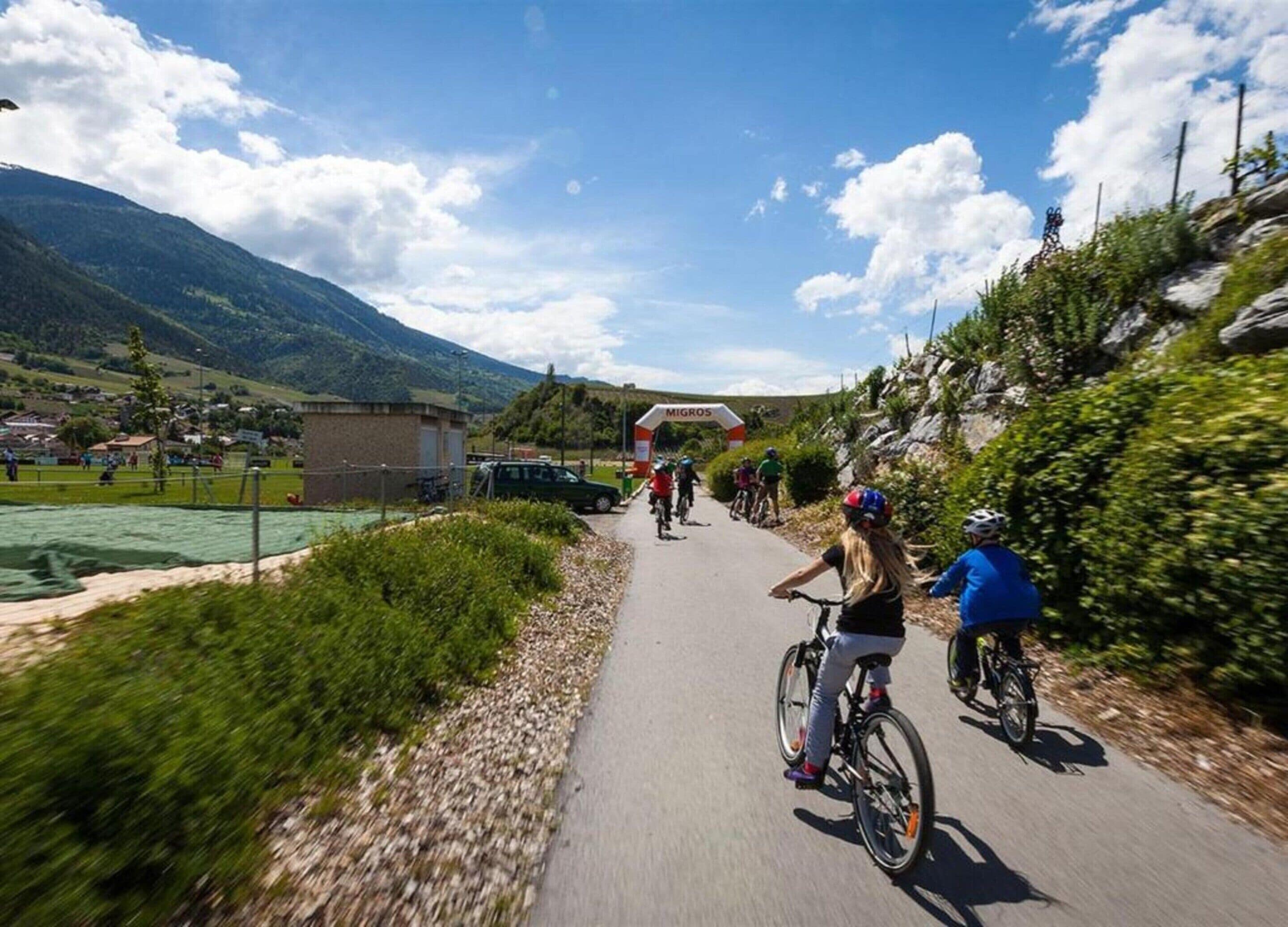 A paved cycling path with cyclists, a sign reading "MIGROS", and a mountainous backdrop.